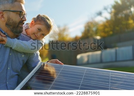 Father with his little daughter catching sun at solar panel,charging at their backyard. Alternative energy, saving resources and sustainable lifestyle concept.