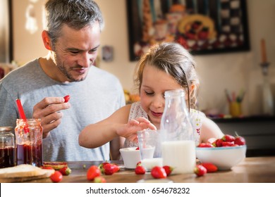 A father and his little girl eating strawberries and sugar in the kitchen. There are a bottle of milk and pot of jam on the table - Shutterstock ID 654678232