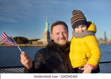 Father And His Little Son Holding The American Flag On The Background Of The Statue Of Liberty. The Concept Of Family Travel