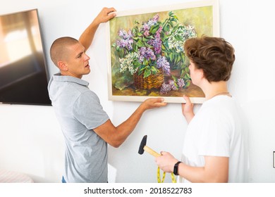 Father And His Son Hanging Picture On Wall. Dad Holding It In Hands And Son Standing With Hammer.