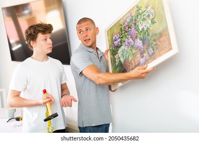 Father And His Son Hanging Picture On Wall. Dad Holding It In Hands And Son Standing With Hammer.