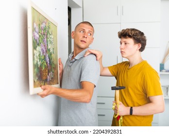 Father And His Son Hanging Picture On Wall. Dad Holding It In Hands And Son Standing With Hammer.