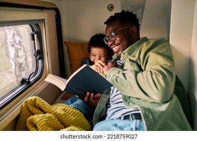 A father and his son smile and read a funny book in a van, while sitting on a bed next to a window.