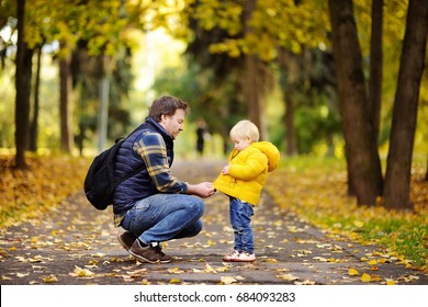 Father And His Toddler Son Walking In Autumn Forest. Family Outdoor Leisure In Park. Man Helping Son Buttoning Zipper On Coat