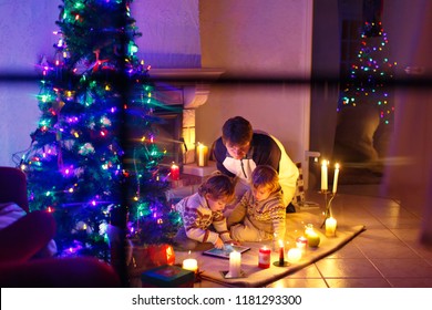 Father And His Two Little Children Sitting By Fireplace Chimney On Christmas Eve Time.