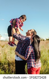 A Father Holding Up His Son, With The Mother At His Side In The Meadow.