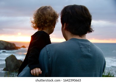 Father Holding His Toddler Daughter As They View The Sunset Over A Pacific Northwest (USA) Beach And Ocean