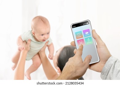 Father Holding Smartphone With Baby Monitor App While Mother Playing With Child