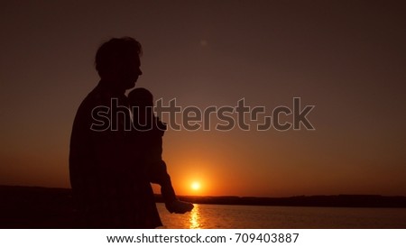 Father holds baby in his arms near water on against red sunset. Slow motion.