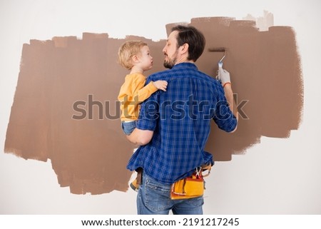 A father holds his little son in his arms and together they draw clouds in brown. The child is wearing an orange blouse, and the man is wearing a blue checkered shirt. The photo is taken from back.