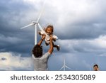 A father joyfully lifts his smiling daughter in the air, with wind turbines and a cloudy sky in the background.