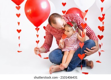 A father kisses his little daughter on Valentine's Day, holding a red ball with a garland of hearts in his hand. Girl sucks a heart-shaped lollipop. Tenderness and the relationship of fathers and kids