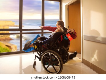 Father Kneeling Next To Disabled Son In Wheelchair Looking Out Hospital Window At Ocean And Cliffs Outdoors. Child Recently Had Head Surgery.