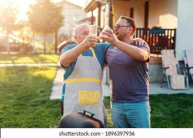 Father In Law And His Son In Law Making A Toast With Beer And Hugging While Standing Next To Grill. Family Gathering Concept.