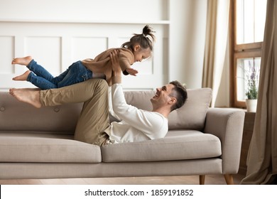 Father Lift Up Daughter While Lying On Couch In Living Room Play Together On Weekend Having Fun Laughing Feels Happy. Little Girl Flying In Air Stretched Arms Like Plane Wings. Family Travel Concept