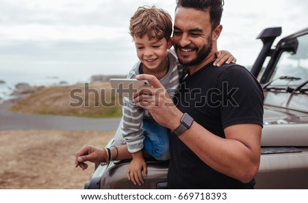Father with little boy using smart phone together during road trip. Young boy with his father sitting at the front of the car.