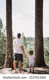 Father and little son holding hands walking in summer pine forest together. Hyper-local travel, family outing. Rear view of man and child hiking in park