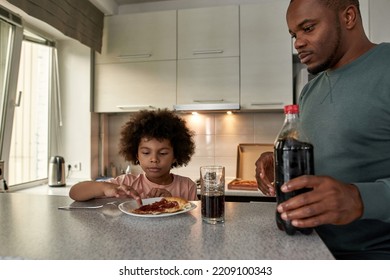 Father Looking At His Little Son Having Lunch Or Dinner With Cola And Pizza At Table At Home Kitchen. Unhealthy Eating. Young Black Family Lifestyle And Relationship. Fatherhood And Parenting