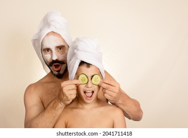 Father looks at the camera while covering his son's eyes with cucumber slices. Caucasian people. Spa day concept, towels on head, family. 
