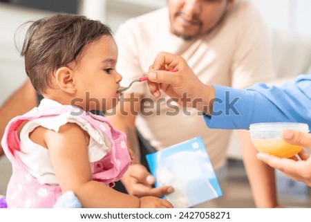 A father looks on with love as his baby girl is being spoon-fed by someone else, a family mealtime scene.