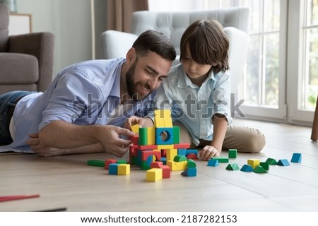 Father lying on warm floor in living room play with preschooler son use wooden multicoloured blocks, construct castle look focused spend pastime together at modern home. Developmental games with