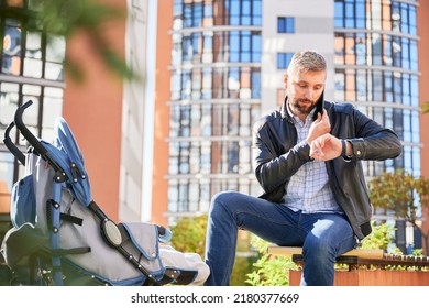 Father Making Business Call, Looking On Watch While Spending Time With Son Outdoors. Front View Of Bearded Man Holding Mobile Phone, Calling, While Baby Sleeping In Pram In Park. Concept Of Working.