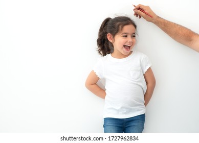 Father Measuring Height Of A Cute Child. Dad Measures The Growth Of Her Child Daughter At A Blank White Wall.