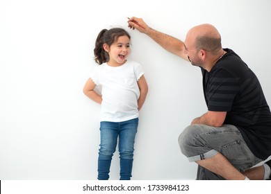 Father Measuring Height Of A Cute Child. Dad Measures The Growth Of Her Child Daughter At A Blank White Wall.