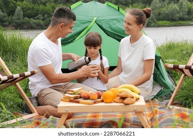 Father mother daughter sitting near tent on the ground having picnic together with delicious fruit and sandwiches man pouring tea from thermos family resting together. - Powered by Shutterstock