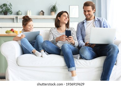 Father, mother and daughter using electronic devices sitting on sofa at living room - Powered by Shutterstock