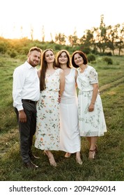 Father And Mother Of Middle Age Tenderly Hug Two Adult Daughters In Nature. Beautiful Family Walks Across The Field At Sunset Of The Day. Parents With Older Children