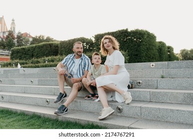 Father, mother, and son are sitting on the steps and staring at the garden of an old European town. Dad is discussing important themes with his smiling family in the park in summer at sunset.