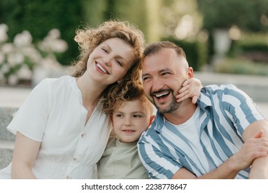 Father, mother, and son are sitting on the steps in the garden of an old European town. A close photo of a laughing family in the park in summer at sunset. - Shutterstock ID 2238778647