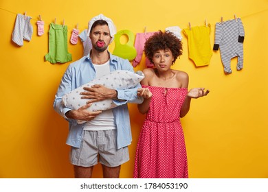 Father, Mother And Their Infant Child. Busy Tired Dad Poses With Baby On Hands, Diaper On Head And Nipple In Mouth, Hesitant Mom Stands Near, Spreads Palms. Parents Care About Newborn. Family Concept