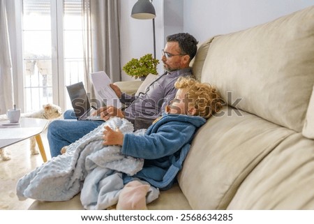 Father multitasking, working remotely on laptop while nurturing sick child, managing professional responsibilities amid home care challenges