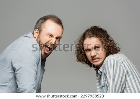 father with opened mouth and disgusted teenage son grimacing and looking at camera isolated on grey