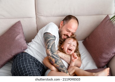 Father Play With Child Kid Girl Spending Time Together. Bald Bearded Caucasian Man In Tattoos Tickles His Tender Little Daughter. Girl Laughs Play With Dad In Pajamas In Bed Morning Routine