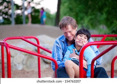 Father Playing With Disabled Son On Merry Go Round At Playground. Child Has Cerebral Palsy. 