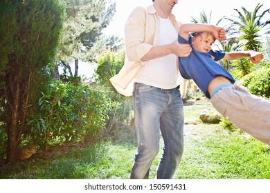 Father Playing With His Child Boy In A Home Garden During A Sunny Day, Picking Him Up And Flying Him Turning Around And Having Fun Together.