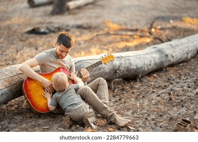 
A father plays the guitar to his son lying on a huge log in the forest in the summer at sunset