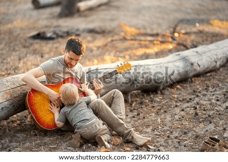 
A father plays the guitar to his son lying on a huge log in the forest in the summer at sunset