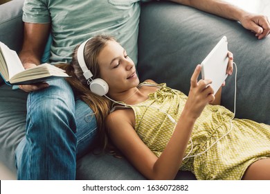 Father And Pre Teen Daughter Playing Tablet And Reading Book While Relaxing Together On Couch In Room At Home. Good Parent And Child Relations.