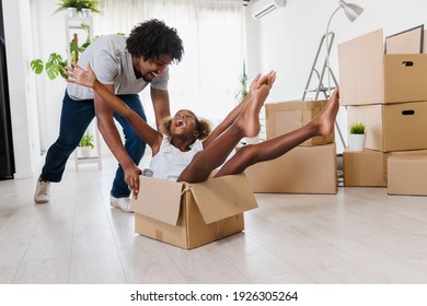 Father Push Cute Little Daughter Sitting Inside Of Carton Box Having Fun Riding In Living Room. Loan Mortgage, Housing Improvement Concept. Cheerful Happy African Family Enjoy Relocation Day.