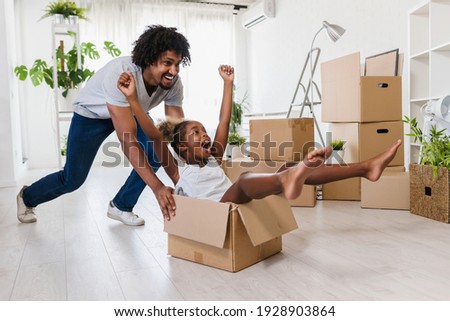 Father push cute little daughter sitting inside of carton box having fun riding in living room. Loan mortgage, housing improvement concept. Cheerful happy african family enjoy relocation day.