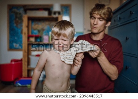 Father putting shirt on little son, changing from pajamas in the morning. Boy is sad, crying.