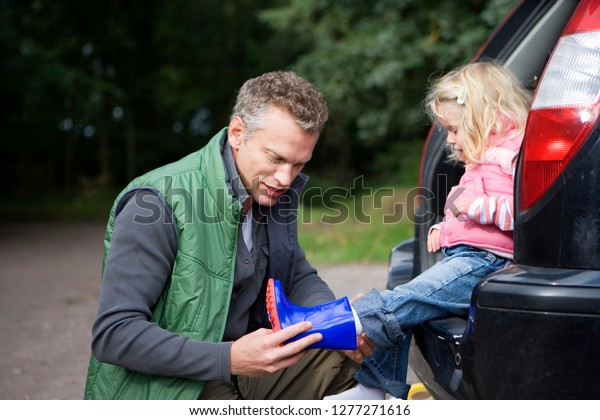 Father putting wellington boot on child\
foot for countryside walk as she sits in car\
trunk