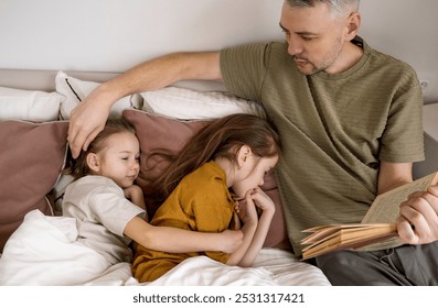 A father is reading a bedtime story to his two daughters who are asleep in bed. He is gently stroking the hair of the daughter closest to him, while the other daughter is resting peacefully.  - Powered by Shutterstock