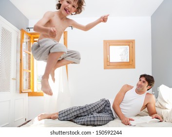 Father reading at book in his bedroom while young son child is jumping up on the mattress, having fun and being energetic during a sunny weekend morning at home.