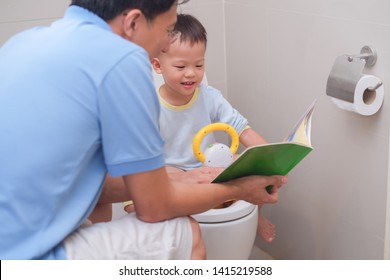 Father Reading Book To Teach Smiling Son To Use Toilet In Bathroom, Cute Little Asian 2 -3  Years Old Toddler Boy Sitting On Toilet With Kid Bathroom Accessory, Happy Potty, Toilet Training Concept