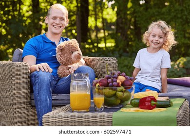 Father Resting With Child In Garden, Sitting On Rattan Furniture